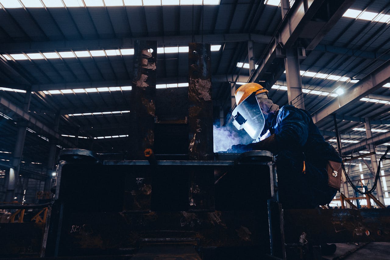 Home A welder wearing protective gear works on metal in a dimly lit industrial workshop.