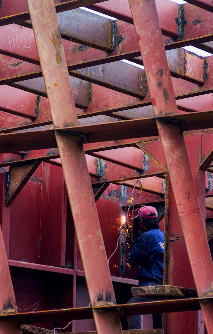 Offerings A welder in action working on a large red metal framework, sparking visible.